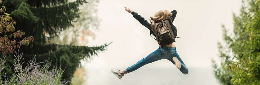 Rapariga feliz com mochila às costas na natureza dá um pulo e aponta para o céu.